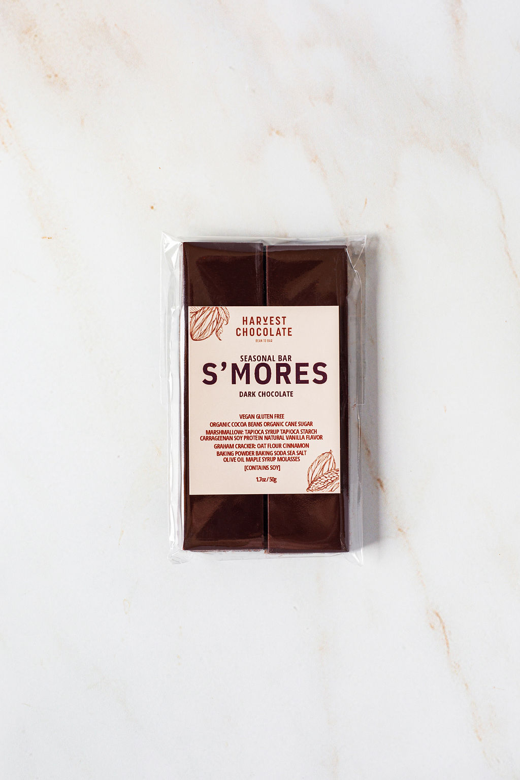 A bar of Harvest Chocolate S'mores is placed on a marble surface. The transparent packaging showcases the vegan and gluten-free chocolate bar inside, with the label detailing the seasonal flavor and featuring illustrations of cocoa beans.