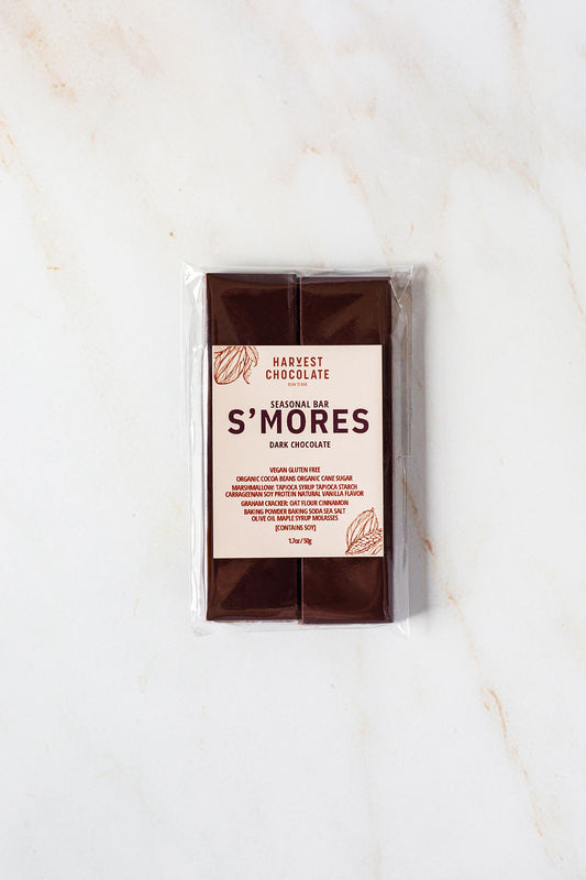 A bar of Harvest Chocolate S'mores is placed on a marble surface. The transparent packaging showcases the vegan and gluten-free chocolate bar inside, with the label detailing the seasonal flavor and featuring illustrations of cocoa beans.