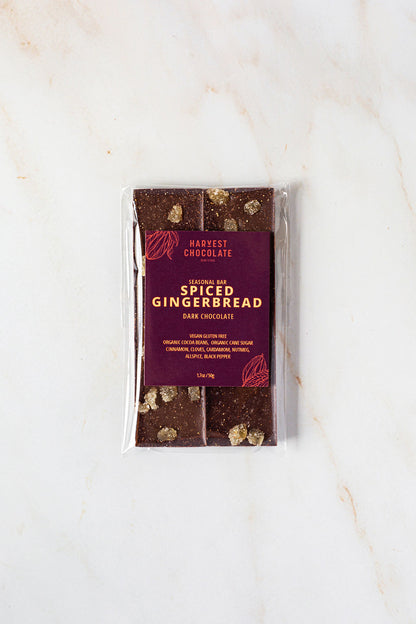 A package of Harvest Chocolate's "Spiced Gingerbread" seasonal bar is placed on a marble surface. Visible pieces of gingerbread, enhanced with organic ginger, can be seen through the transparent wrapping. The holiday treat's packaging includes ingredients and nutritional information.