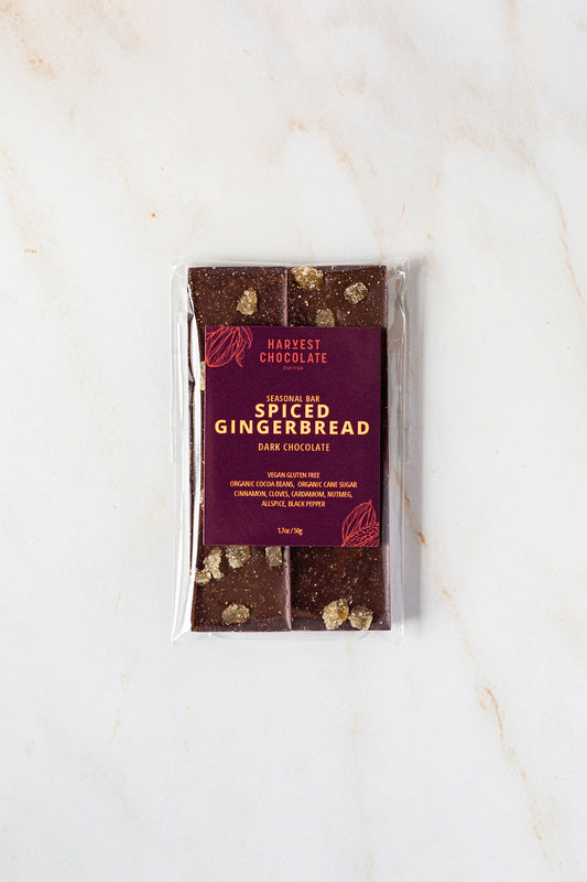 A package of Harvest Chocolate's "Spiced Gingerbread" seasonal bar is placed on a marble surface. Visible pieces of gingerbread, enhanced with organic ginger, can be seen through the transparent wrapping. The holiday treat's packaging includes ingredients and nutritional information.