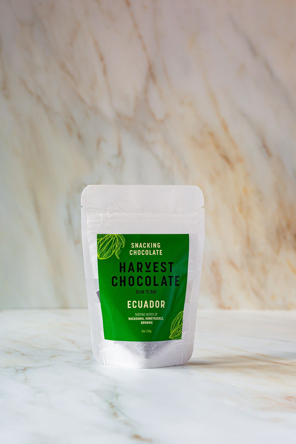 A small, white, resealable pouch stands upright on a marble surface. The front green label reads "Ecuador Snacking Chocolate, Harvest Chocolate." Leaf illustrations adorn the corners. Crafted with organic ingredients and perfect for vegan diets.