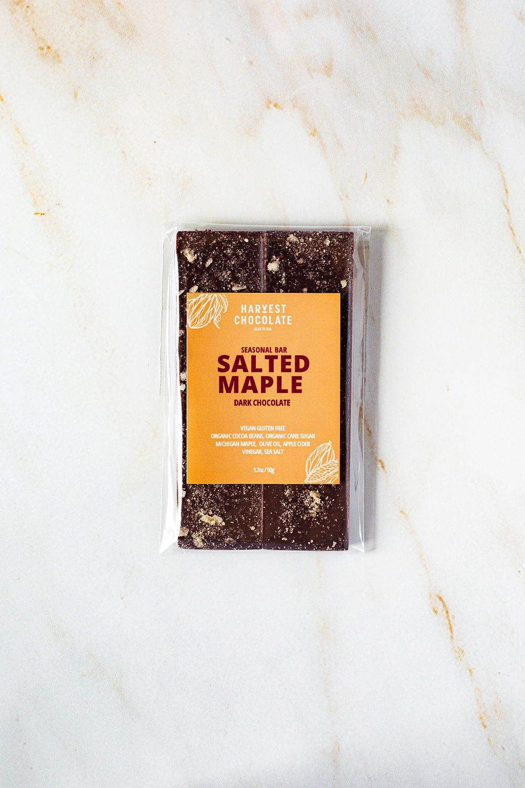 A package of Harvest Chocolate's Salted Maple bar, a seasonal treat of dark chocolate infused with Michigan maple syrup, is housed in transparent packaging with an orange label showcasing the flavor and product details. The bar elegantly rests on a light-colored marble surface.