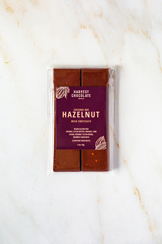 A Hazelnut Milk Chocolate bar, labeled "Harvest Chocolate," is wrapped in clear packaging and rests on a marble background. The bar is vegan, gluten-free, and made with locally grown Hazelnuts.