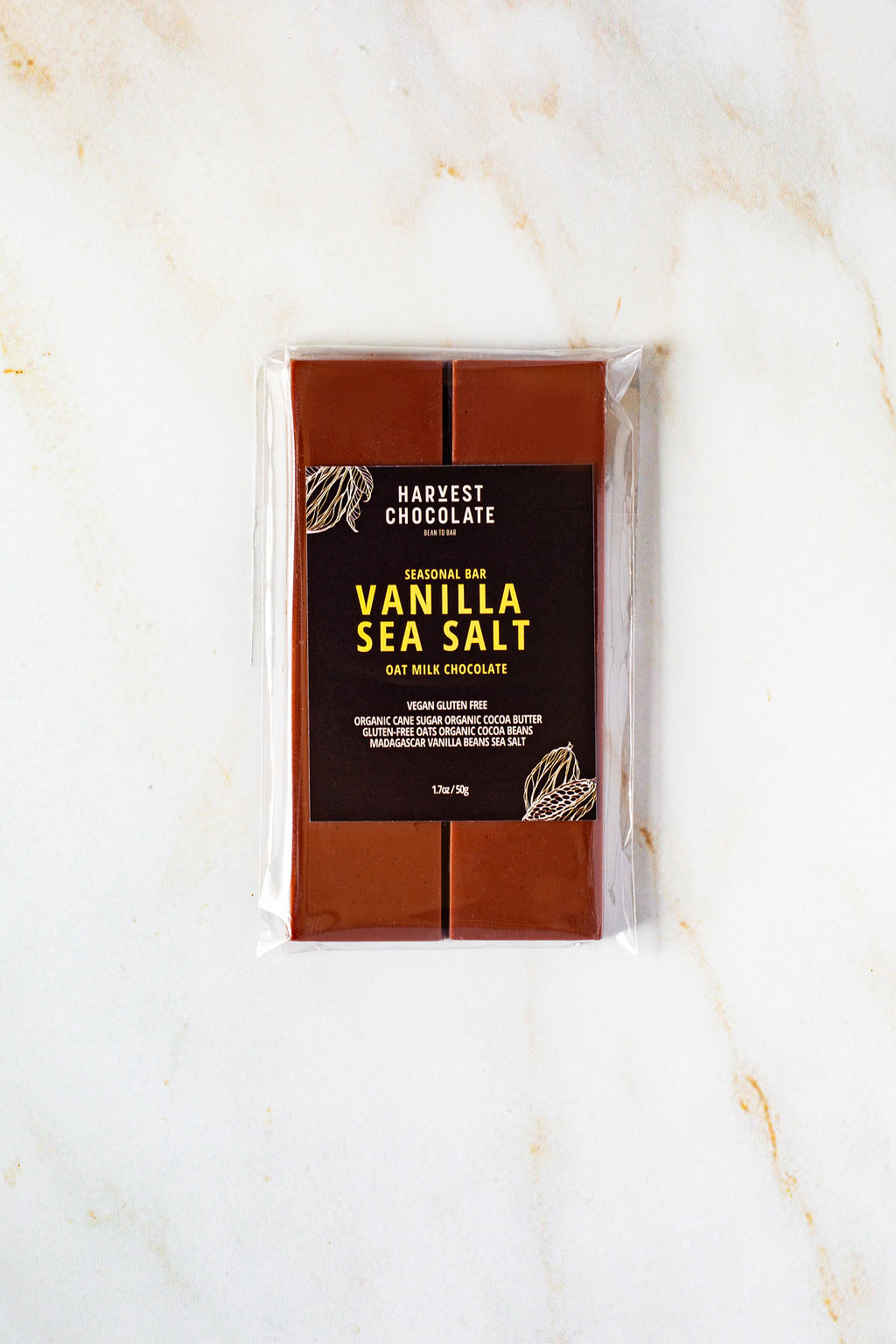 A packaged Harvest Chocolate bar rests on a white marbled surface. The label announces "Vanilla Bean Sea Salt, Oat Milk Chocolate, Seasonal Bar." It is described as "Vegan, Gluten-Free, Organic, with 60% Cocoa Butter and Contains Coconut." The bar has a weight of 1.75 oz (50g).