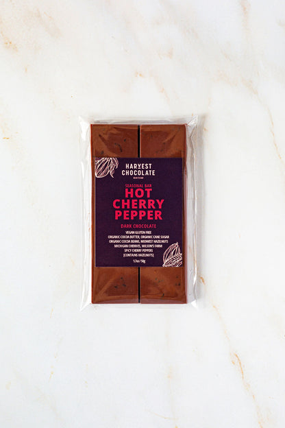 A Harvest Chocolate bar named "Hot Cherry Pepper." The dark chocolate bar comes in clear plastic packaging, showcasing its segmented design. The label highlights its status as seasonal and made with organic ingredients, Michigan cherries, and natural hot cherry pepper flavors.