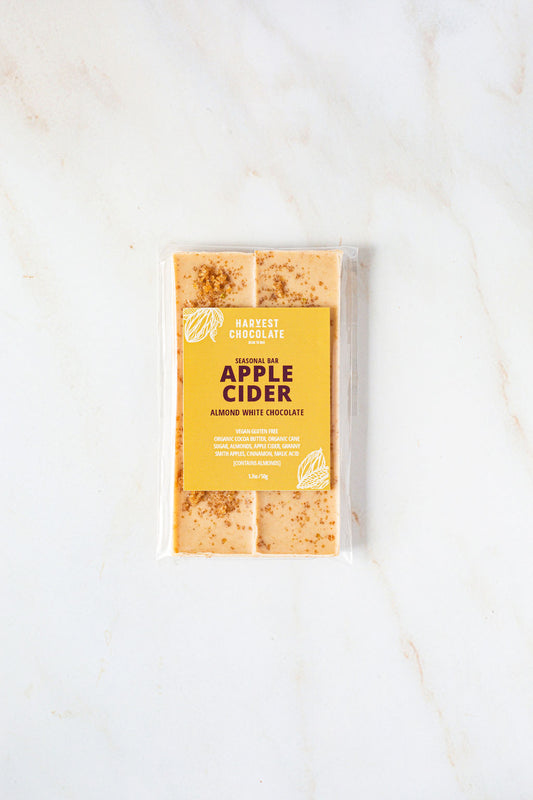 A packaged Apple Cider bar from Harvest Chocolate is shown against a light marble background. The label features a yellow design with the brand name and product details. Made with organic apples, the almond white chocolate bar has a slightly textured surface.