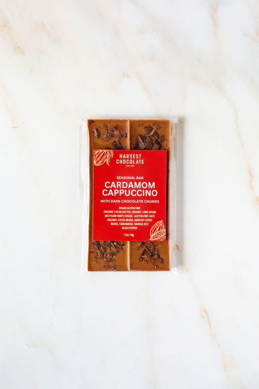 A packaged chocolate bar from Harvest Chocolate is placed on a light surface. The red label clearly displays the flavor: "Cardamom Cappuccino." The packaging is transparent, showcasing the chocolate and dark chunks inside. This treat is ideal for coffee aficionados, particularly fans of Madcap Coffee.