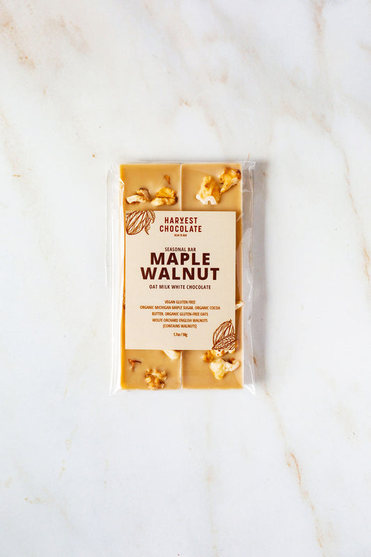 A packaged bar of Harvest Chocolate labeled "Maple Walnut" rests on a marble surface. The transparent wrapper reveals pieces of English walnut embedded in the chocolate. The packaging includes ingredient and nutritional information.