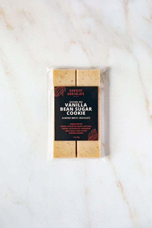 A packaged chocolate bar labeled "Vanilla Bean Sugar Cookie" from Harvest Chocolate lies on a light marble surface. The label reads "Almond White Chocolate," highlighting its organic ingredients and lactose-free status. Segmented into square pieces, this treat is a perfect blend of flavors.