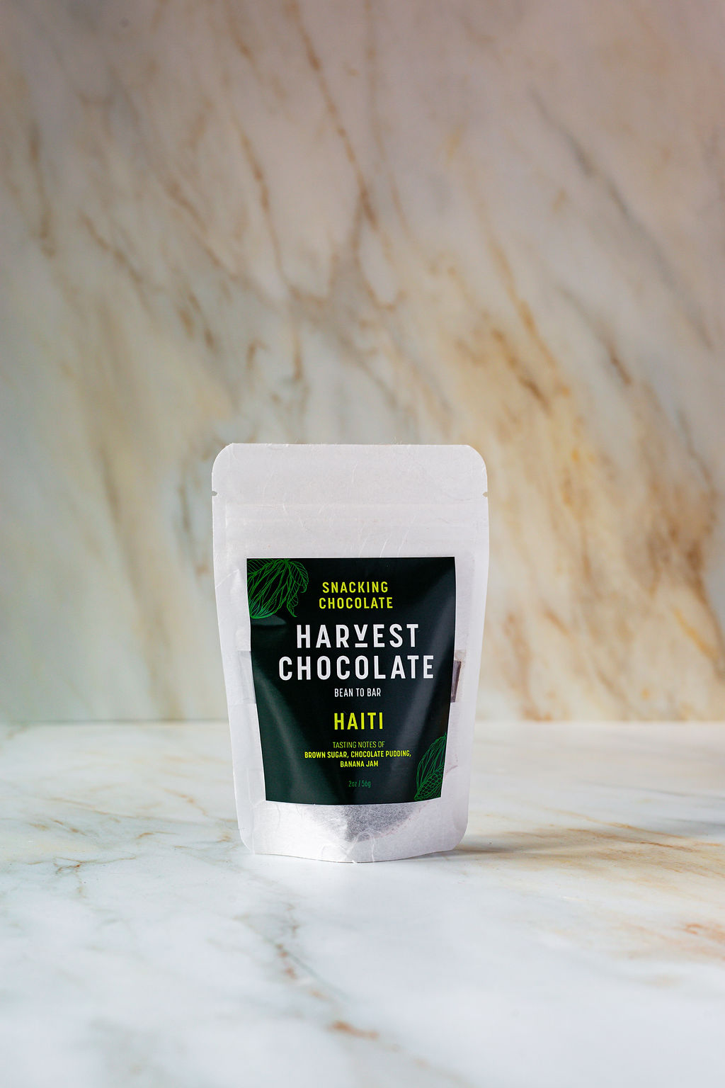 A small, white, resealable bag of Haiti Snacking Chocolate stands on a marbled surface. The packaging has a black label with green accents and the text "Harvest Chocolate - Bean to Bar - Haiti" printed on it, highlighting its commitment to being organic dark chocolate.