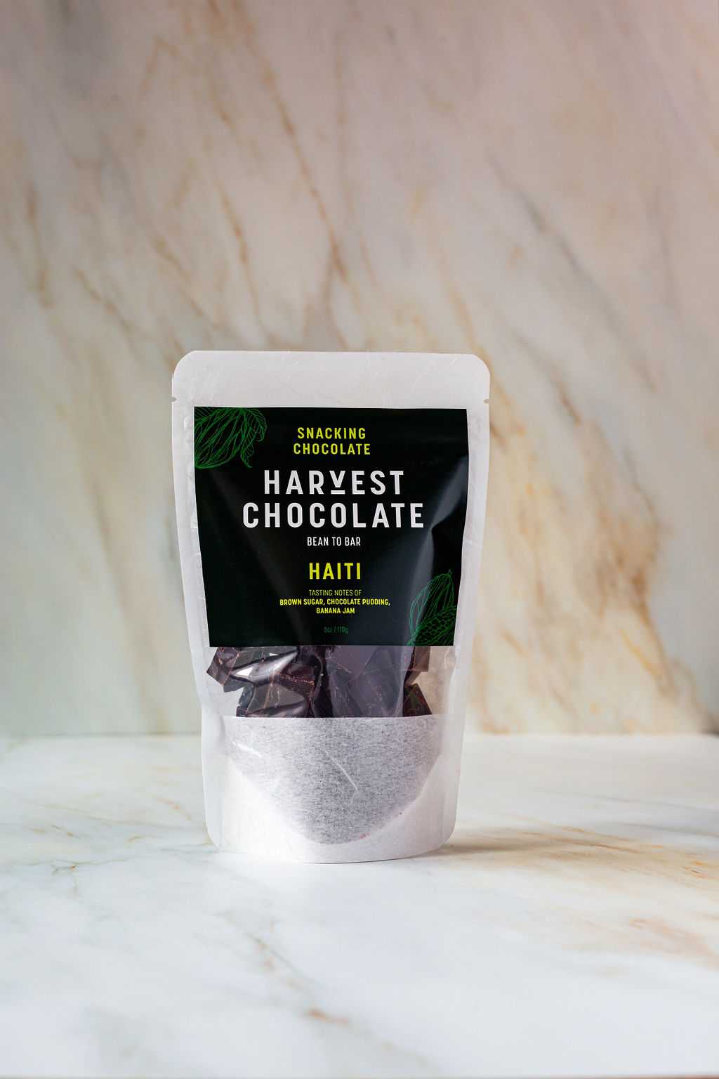 A white, resealable pouch with a black label displaying the text "Haiti Snacking Chocolate - Harvest Chocolate" rests on a light marble surface. The pouch contains organic dark chocolate pieces visible through a small transparent window at the bottom.