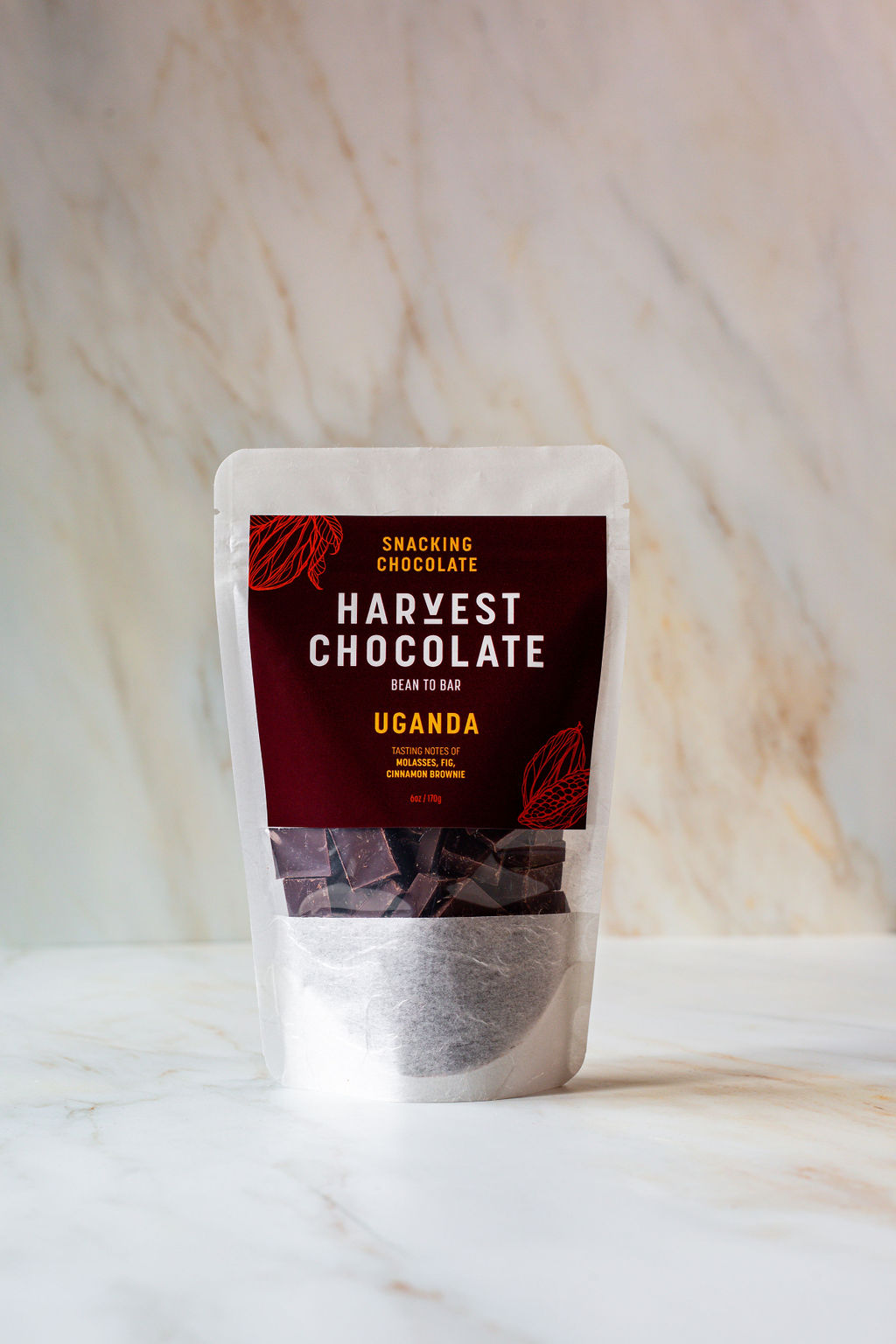 A pouch of Uganda Snacking Chocolate by Harvest Chocolate, featuring a maroon label with white text that reads "Harvest Chocolate Bean to Bar Uganda," is set against a marble background. Inside the transparent lower part of the pouch, the vegan dark chocolate pieces promise a rich fudgy flavor.