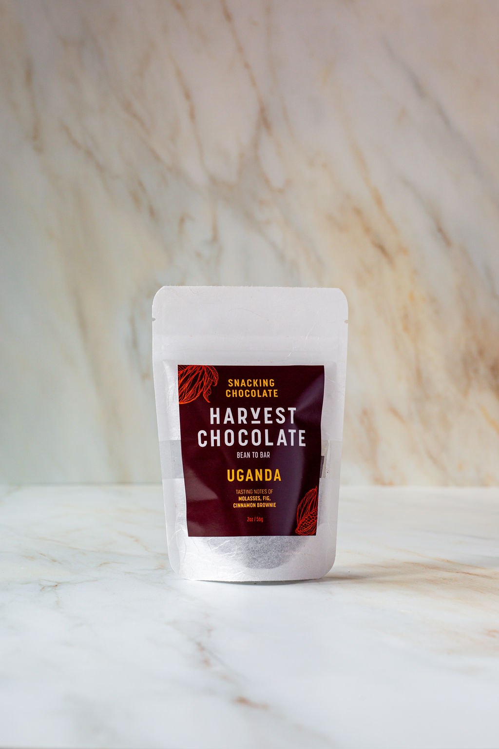 A white pouch of "Uganda Snacking Chocolate" by Harvest Chocolate is centered in the image. The dark brown label reads, "Snacking Chocolate: Uganda Snacking Chocolate, Bean to Bar, Dark Chocolate with Cocoa Nibs and Sea Salt." Made from organic chocolate, this vegan dark chocolate offers a rich fudgy flavor. The background is a light marbled surface.