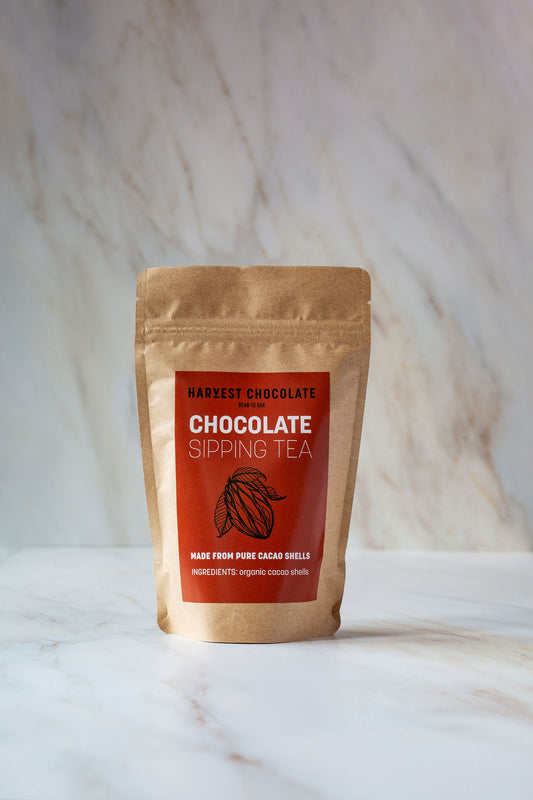 A brown paper bag of "Harvest Chocolate Chocolate Tea" is placed on a marble surface. The packaging has a red label featuring an illustration of cacao and text stating "Made from pure organic cocoa bean shells" and "Ingredients: organic cacao shells.