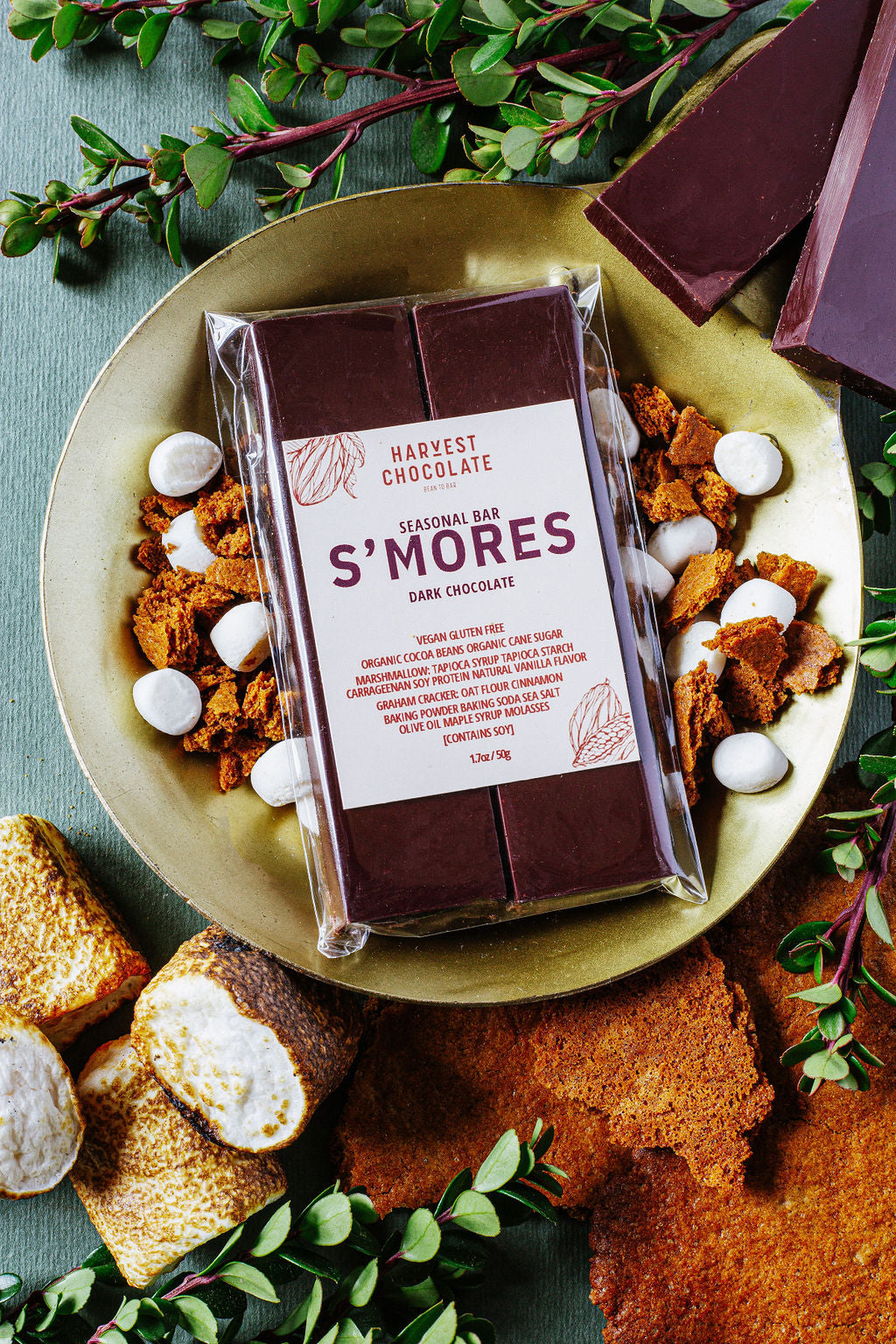 A gold plate holds a Harvest Chocolate S'mores bar wrapped in plastic. The plate is surrounded by crushed graham crackers, mini marshmallows, and chocolate chunks, while toasted marshmallows and greenery are arranged around it.