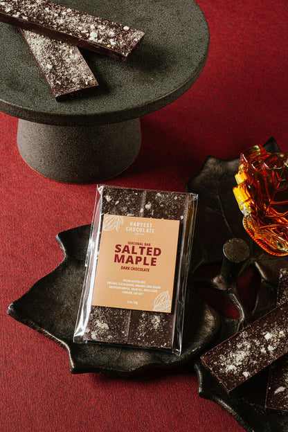 A Harvest Chocolate Salted Maple bar is elegantly displayed on a dark platter with more chocolate pieces. A leaf-shaped plate and a small glass of maple syrup add charm to the scene, set against a rich red background.