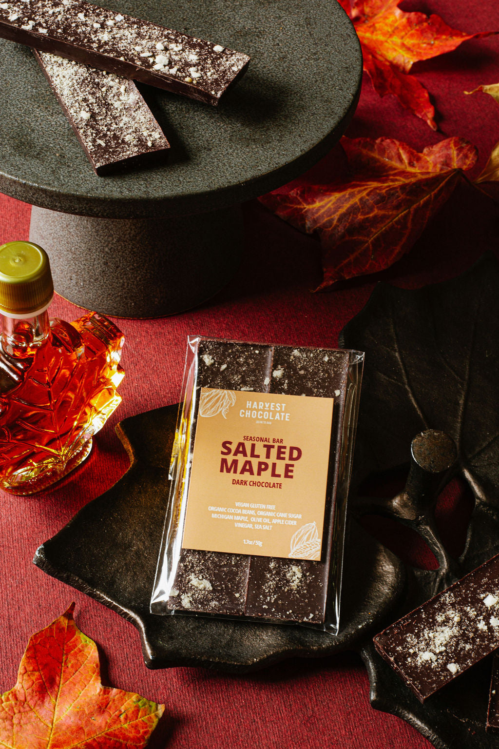 A packaged bar of Harvest Chocolate's Salted Maple chocolate is displayed on a black leaf-shaped platter. Maple leaves and a bottle of Michigan maple syrup are arranged around it on a red surface, with square pieces of chocolate scattered nearby.