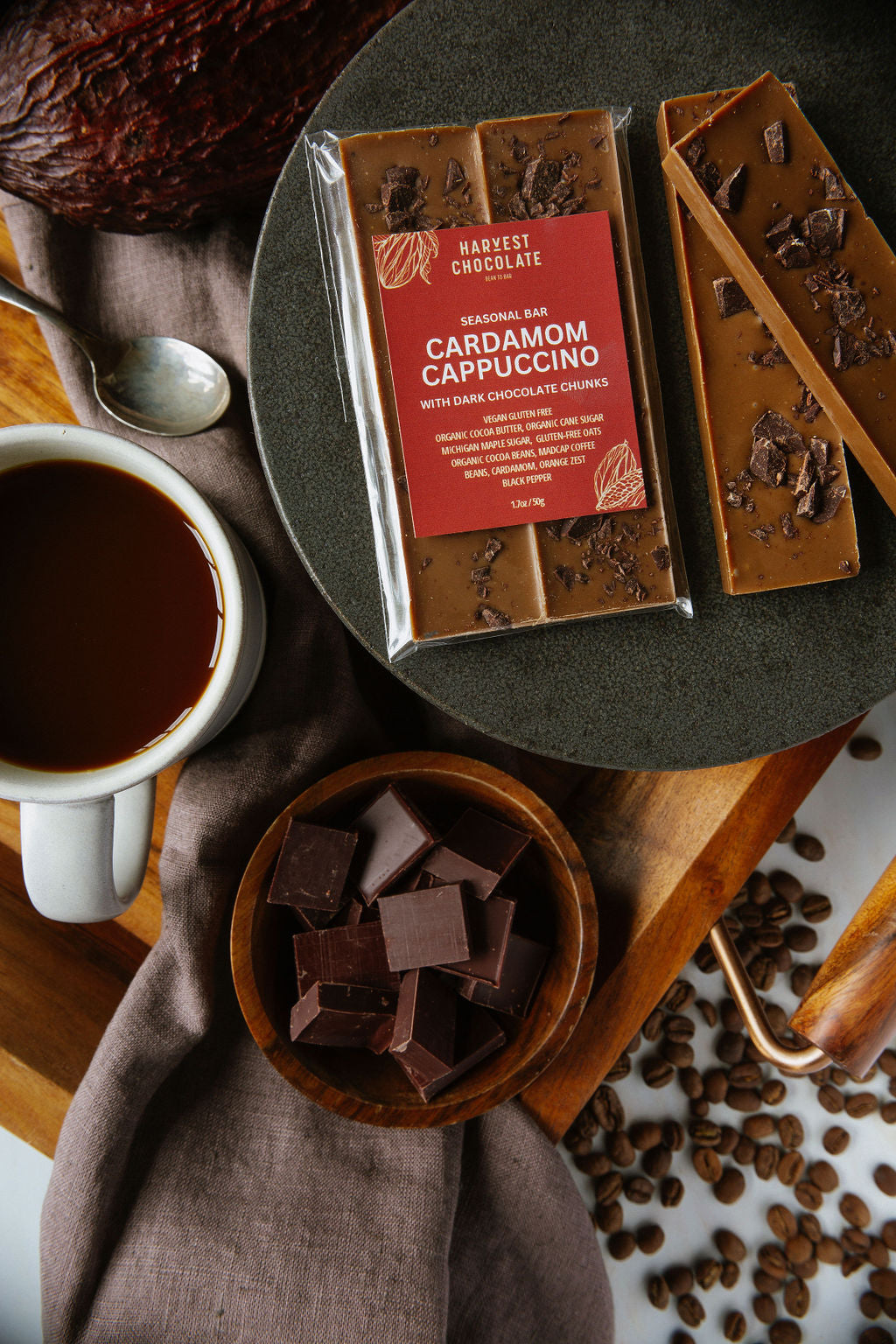 An arrangement of Harvest Chocolate's Cardamom Cappuccino bars, one unwrapped, on a dark plate beside a white coffee cup filled with Madcap Coffee, and a wooden bowl containing square oat white chocolate pieces. Coffee beans are scattered on a wooden surface.