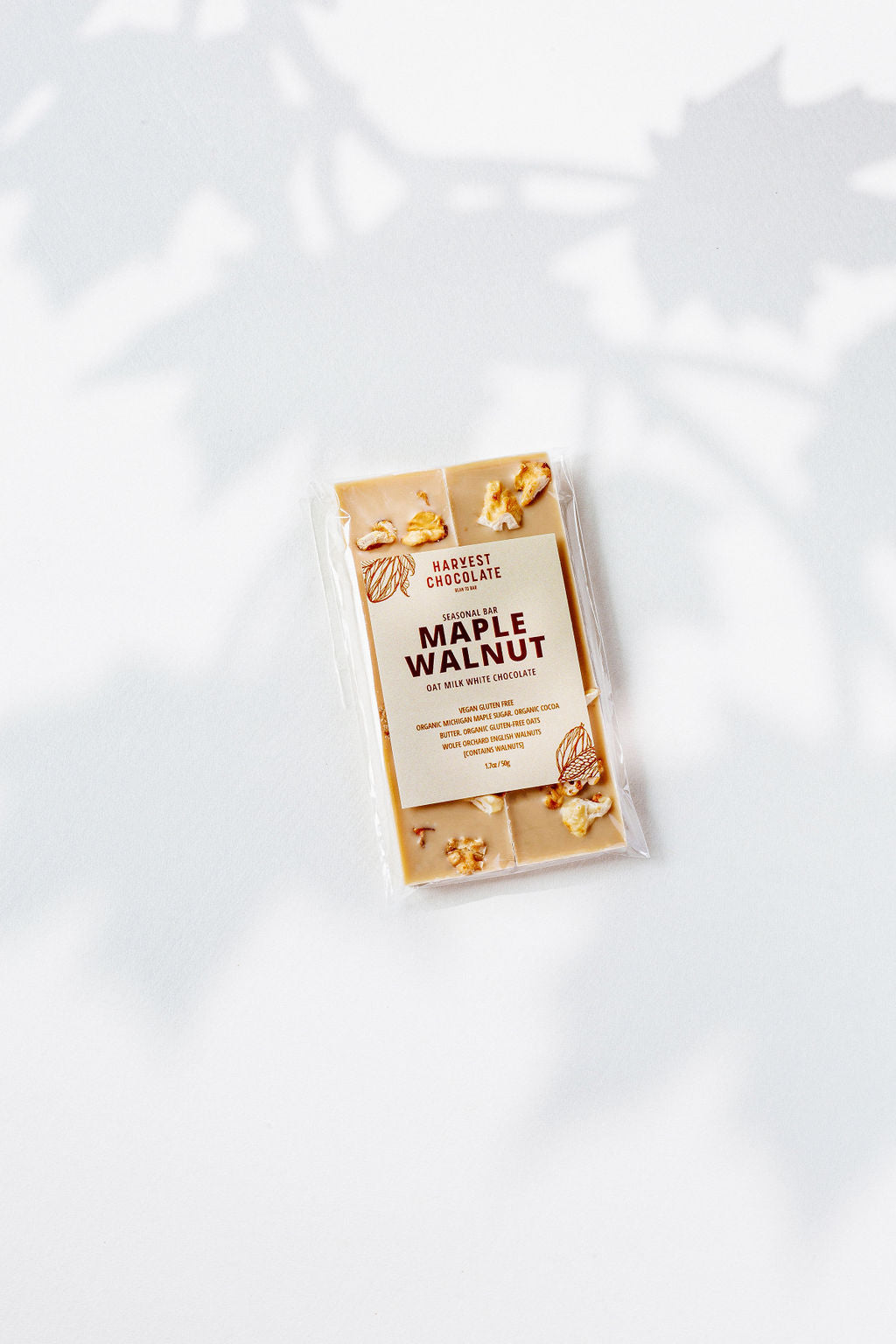 A Harvest Chocolate Maple Walnut bar wrapped in clear packaging lies on a light surface. The package shows the brand and flavor made with English walnuts. Soft leaf shadows in the background enhance this elegant treat.