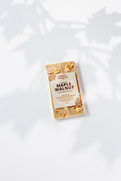 A Harvest Chocolate Maple Walnut bar wrapped in clear packaging lies on a light surface. The package shows the brand and flavor made with English walnuts. Soft leaf shadows in the background enhance this elegant treat.