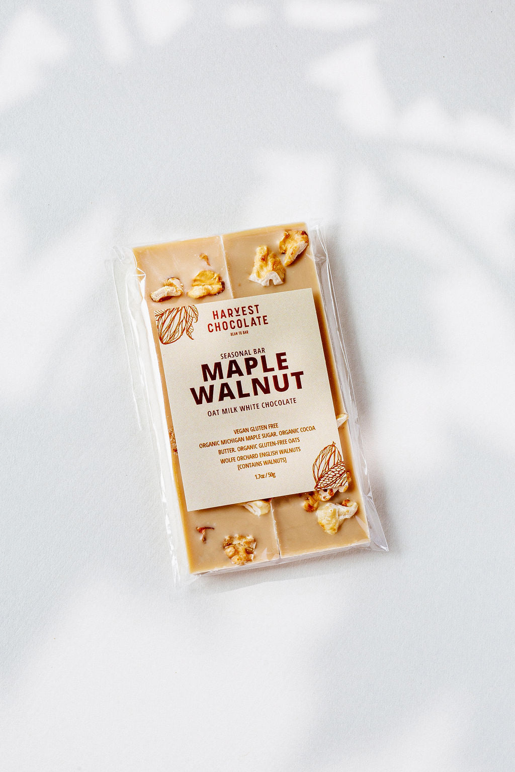 A Maple Walnut oat milk white chocolate bar by Harvest Chocolate rests on a white surface, highlighting its gluten-free and organic ingredients. Enhanced with Michigan maple sugar, it features pieces of English walnut.