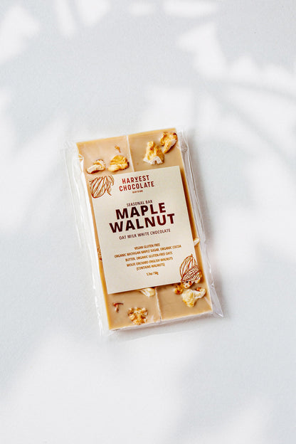 A Maple Walnut oat milk white chocolate bar by Harvest Chocolate rests on a white surface, highlighting its gluten-free and organic ingredients. Enhanced with Michigan maple sugar, it features pieces of English walnut.