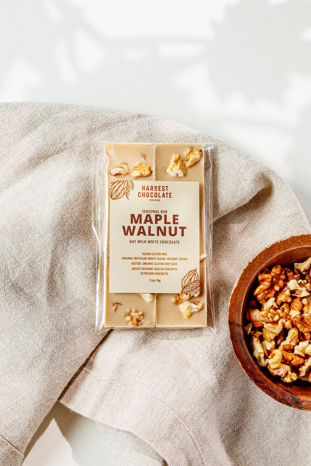 A package of Maple Walnut from Harvest Chocolate rests on a beige fabric, displaying visible walnut pieces. Beside it, there's a wooden bowl filled with loose English walnut pieces.