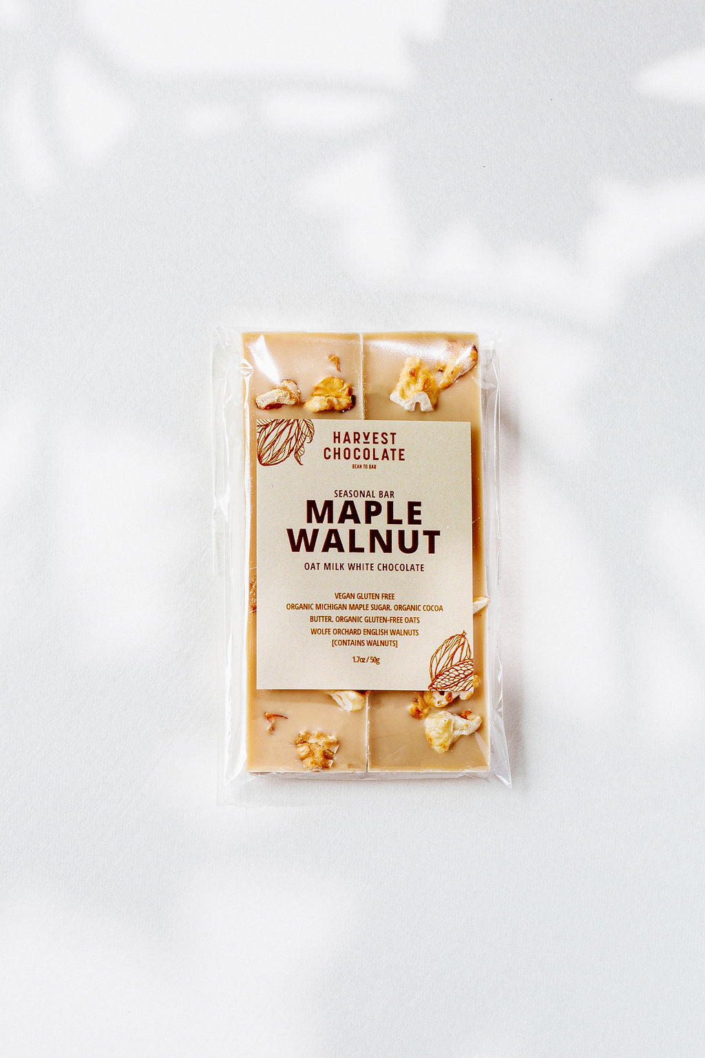 The image shows a package of Harvest Chocolate's "Maple Walnut" oat milk white chocolate with Michigan maple sugar and English walnuts, wrapped in clear packaging and labeled with ingredients like organic cocoa butter.