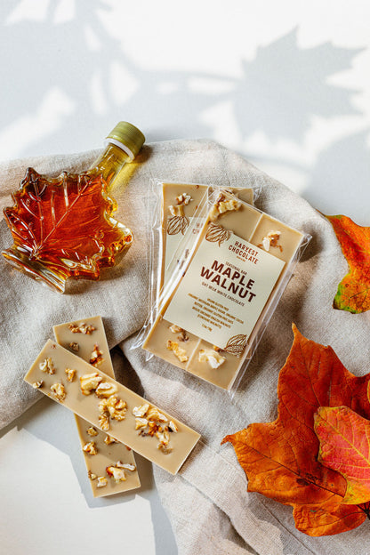 A flat lay image features a maple-shaped bottle of Michigan maple sugar syrup, a wrapped Harvest Chocolate Maple Walnut bar, two unwrapped pieces of the chocolate with visible English walnuts, and several orange and red autumn leaves placed on a neutral fabric background.