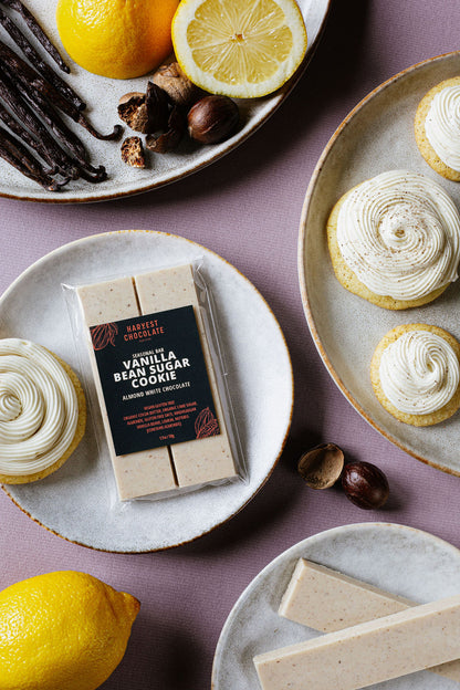 A bar of Harvest Chocolate's Vanilla Bean Sugar Cookie is placed on a plate adorned with piped white chocolate decorations, surrounded by lemons, vanilla beans, and spices on a purple surface, evocative of a sugar cookie. Additional plates with piped decorations are nearby.