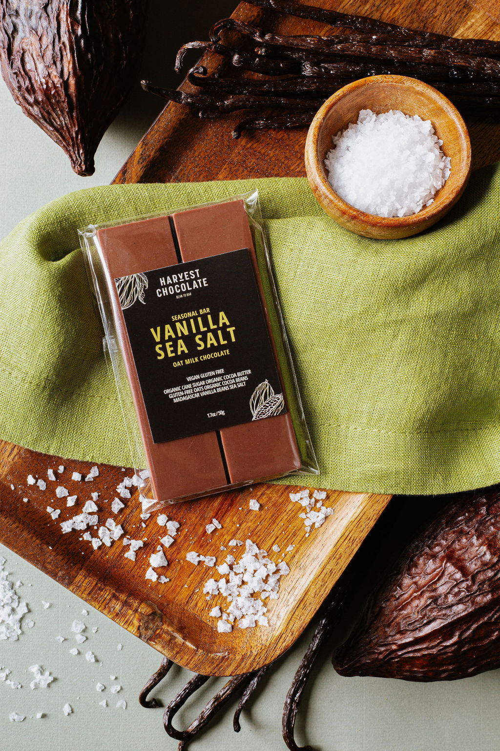 A chocolate bar labeled "Vanilla Bean Sea Salt" from Harvest Chocolate sits on a wooden board with a green cloth. Surrounding the oat milk chocolate bar are vanilla beans, a bowl of sea salt, cacao pods, and scattered salt crystals. The chocolate bar is wrapped in clear plastic.