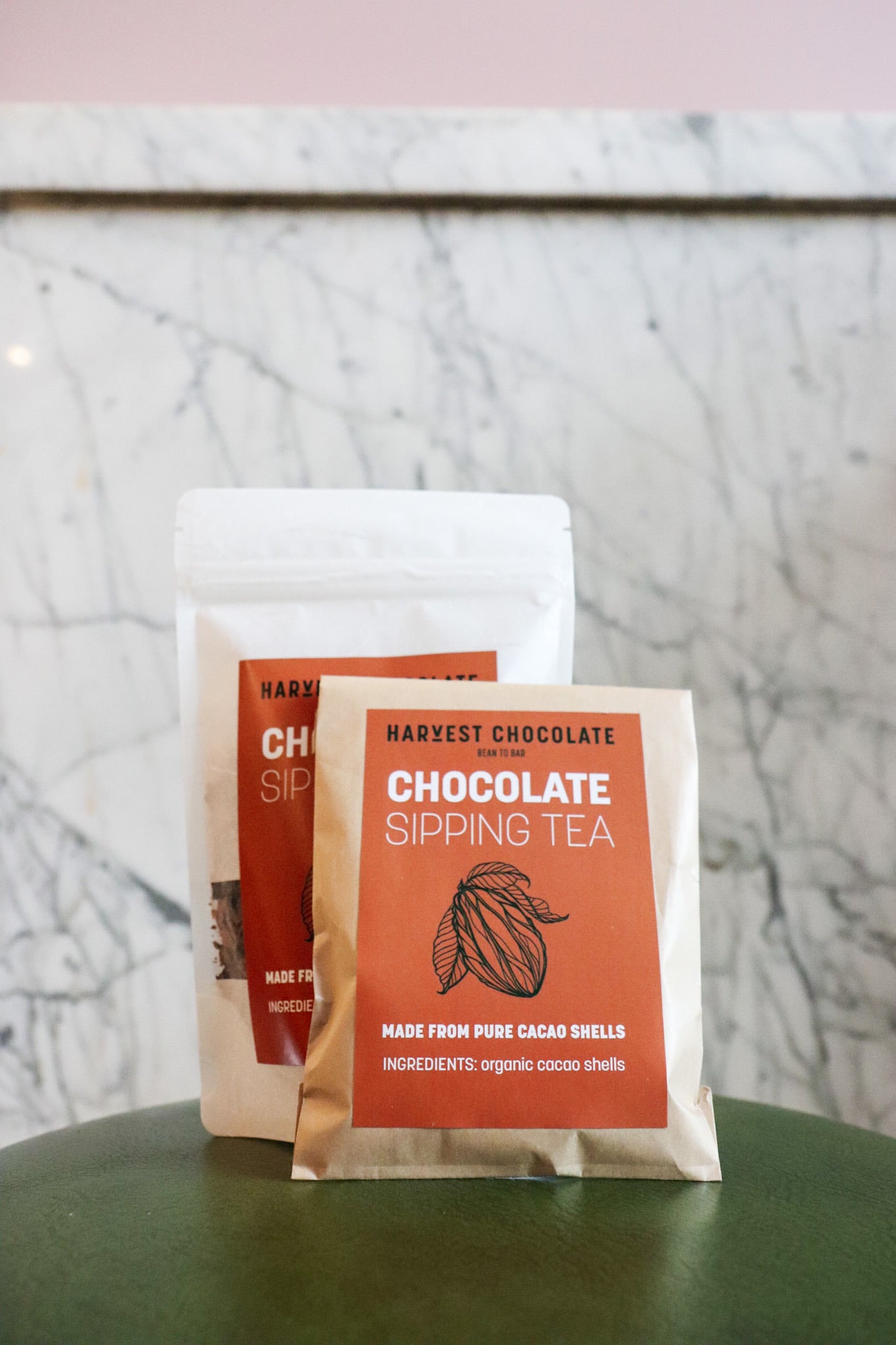 Two packages of Harvest Chocolate Chocolate Tea on a green surface with a marble wall in the background. The packages are labeled and list ingredients including organic cocoa shells.