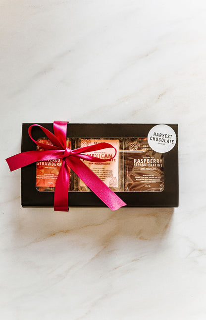 A black rectangular Valentine Trio Box by Harvest Chocolate, adorned with a pink ribbon and white logo sticker, holds three exquisite chocolates: Chocolate Covered Strawberry, Mexican Hot Chocolate, and Raspberry Sesame Praline. Displayed elegantly on a marble surface.