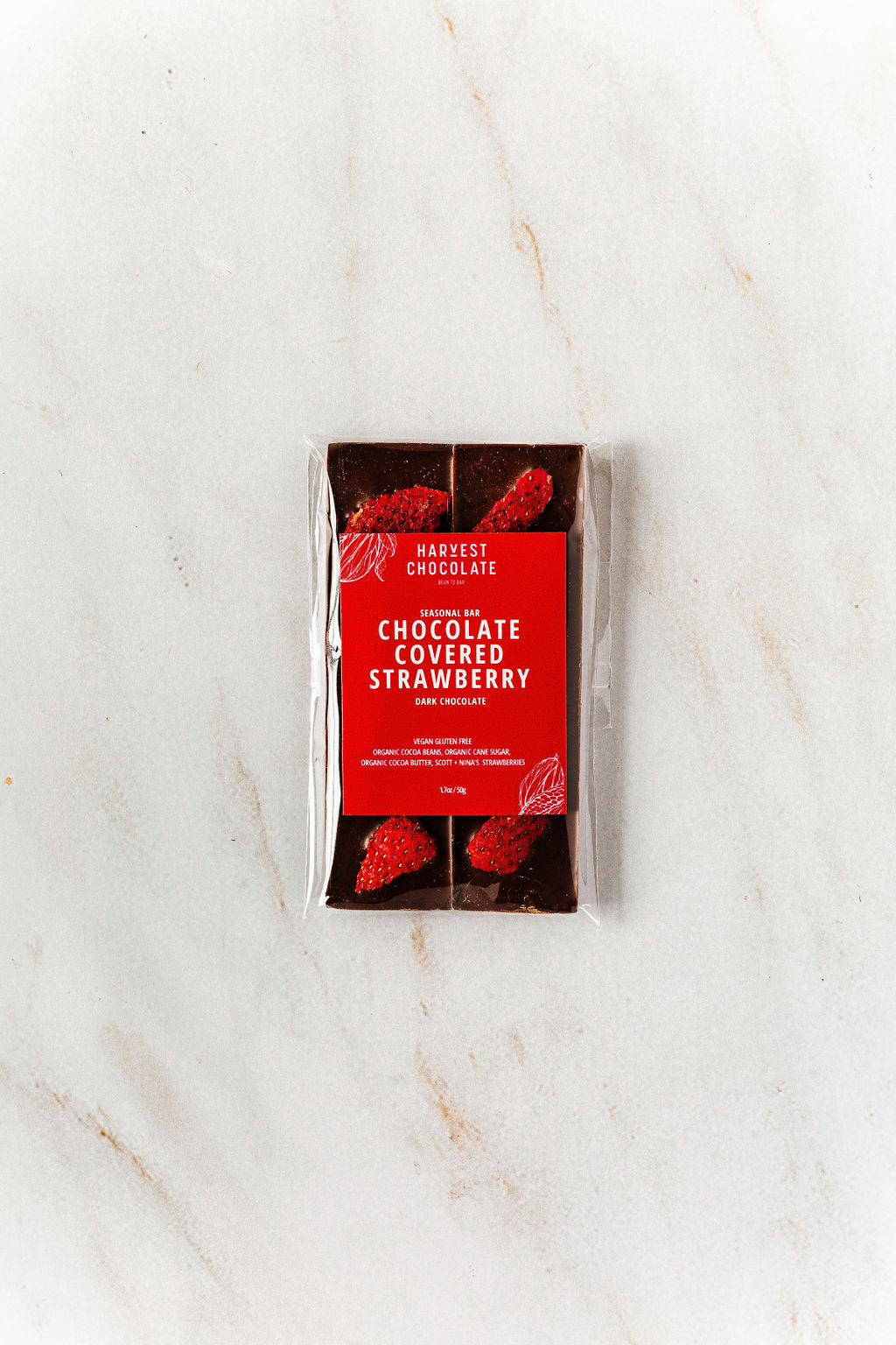 A Harvest Chocolate "Chocolate Covered Strawberry" bar, made with Ugandan cocoa beans, lies on a marble surface. Its transparent packaging showcases chunks with red fruit pieces inside, while a red label with white text stands prominently in the center.