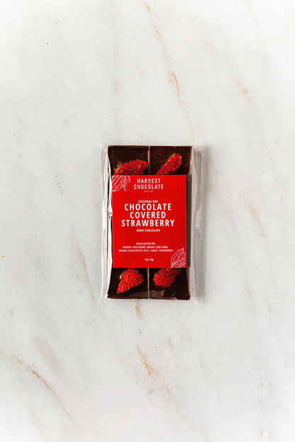 A Harvest Chocolate "Chocolate Covered Strawberry" bar, made with Ugandan cocoa beans, lies on a marble surface. Its transparent packaging showcases chunks with red fruit pieces inside, while a red label with white text stands prominently in the center.