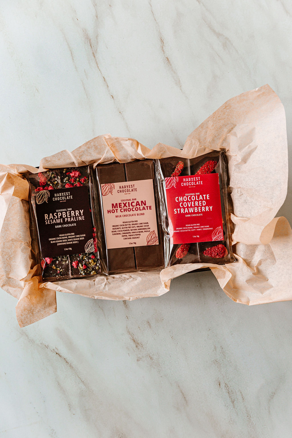 Harvest Chocolate's Valentine Trio Box displays three chocolate bars—Raspberry Sesame Praline, Mexican Hot Chocolate, and Chocolate Covered Strawberry—elegantly wrapped with in a box with a ribbon.