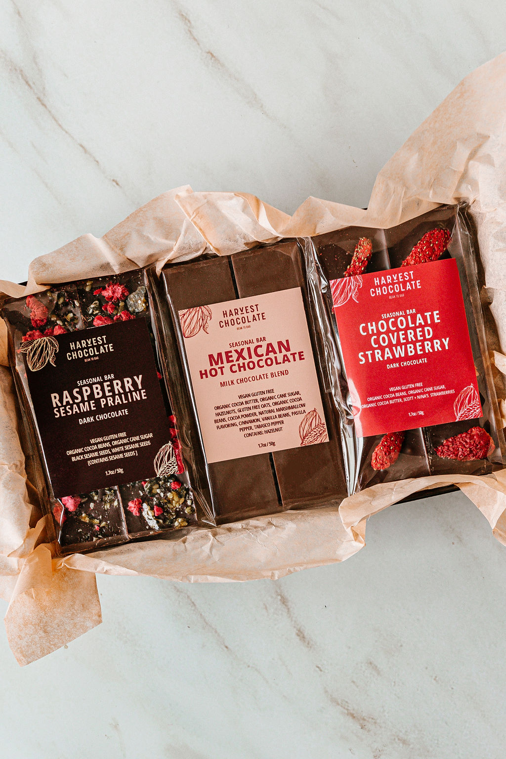 The Valentine Trio Box from Harvest Chocolate includes Raspberry Sesame Praline, Mexican Hot Chocolate, and Chocolate Covered Strawberry bars, all gift-wrapped in a box, pictured against a light marbled surface.