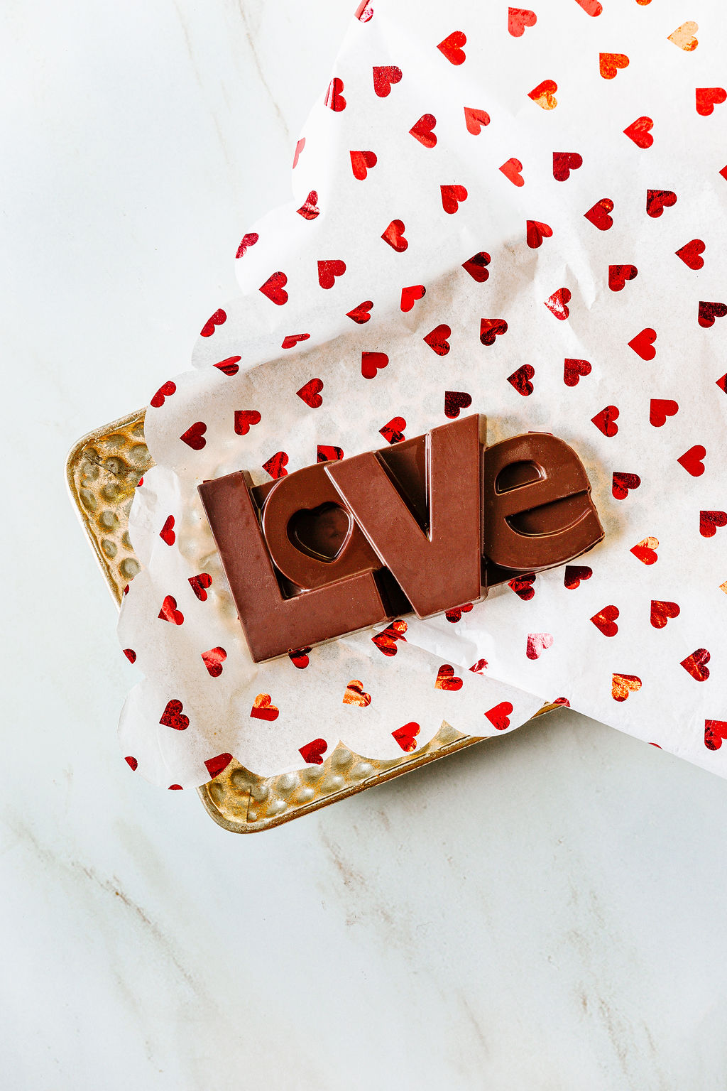 The LOVE Dark Chocolate Bar by Harvest Chocolate, crafted from organic cocoa beans and shaped into "love," rests on heart-patterned tissue paper that partly covers a textured gold tray on a marble surface.