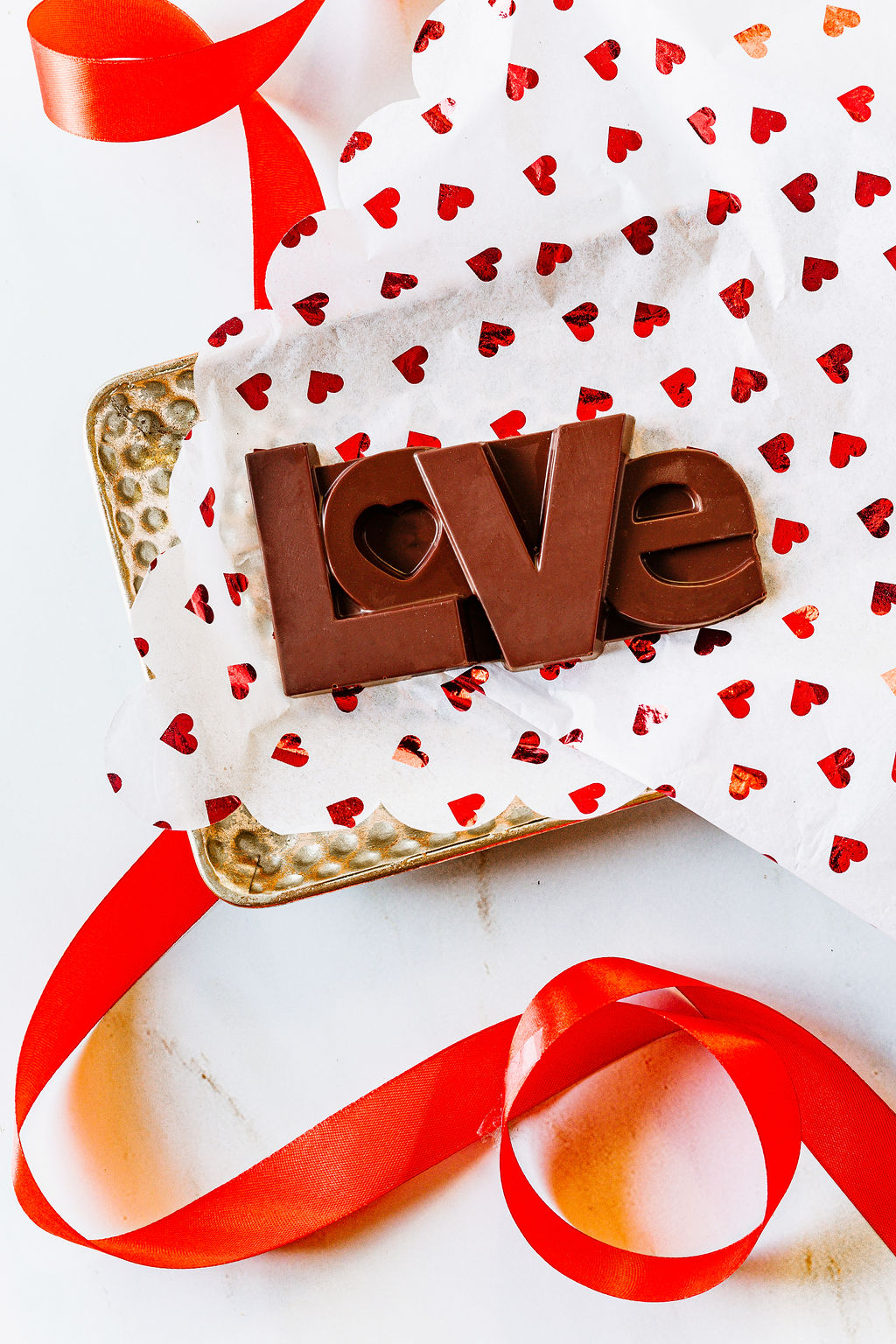 The LOVE Dark Chocolate Bar by Harvest Chocolate features letters spelling "LOVE" crafted from dark chocolate, resting on heart-patterned paper in a gold tray. A red ribbon artistically wraps the tray on a white surface, capturing elegance and sweetness in every detail.