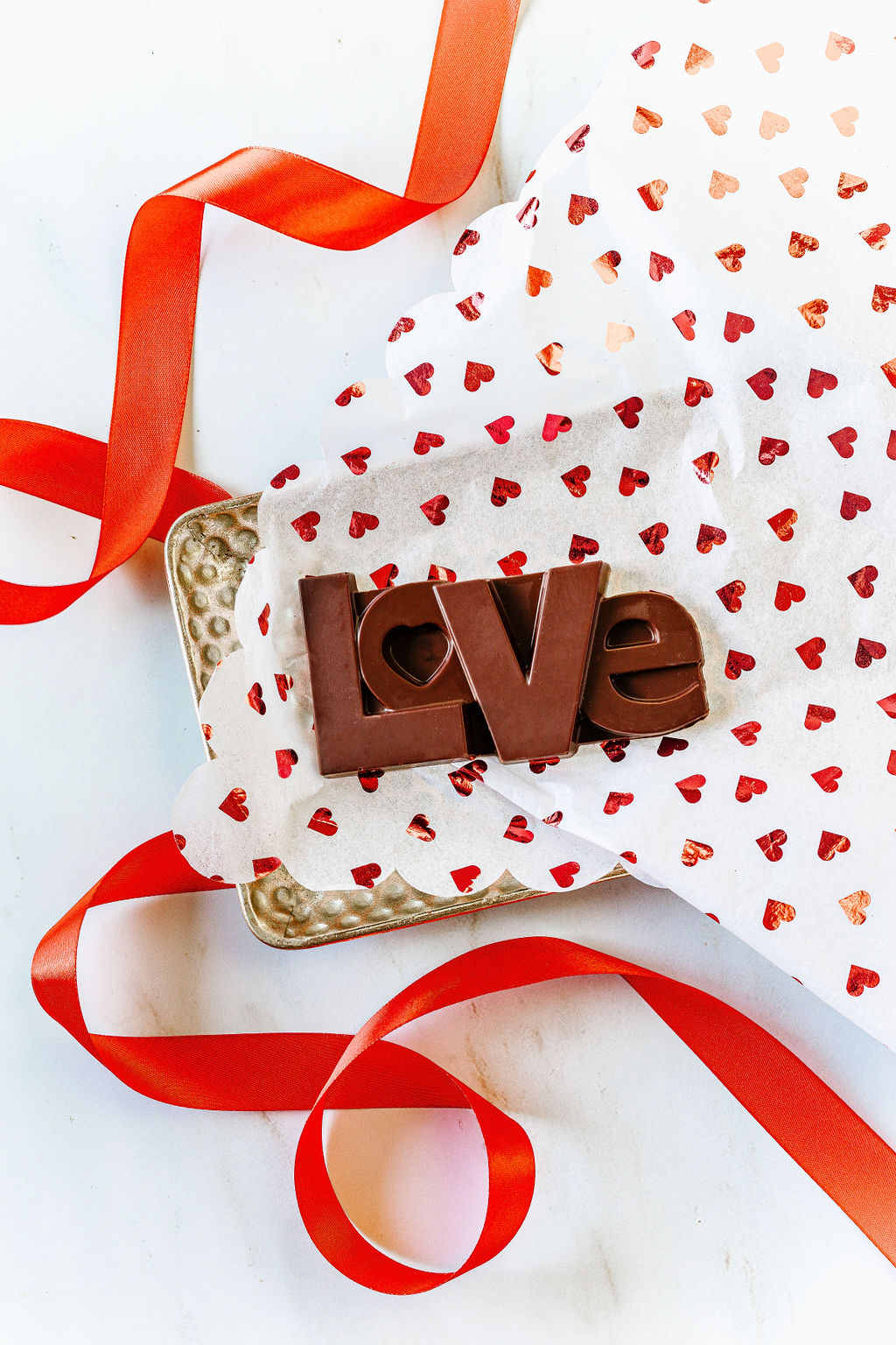 The Harvest Chocolate LOVE Dark Chocolate Bar, made from rich Ecuadorian cocoa beans, is displayed on heart-patterned paper inside a gold tray with a red ribbon artistically enhancing its festive look.