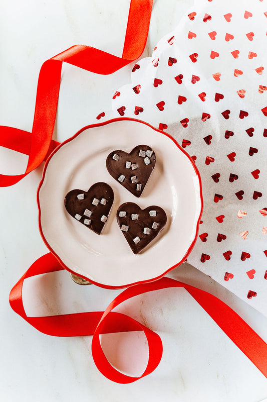 A white plate with red trim holds three Cherry Jam Hearts by Harvest Chocolate, crafted from rich Ugandan cocoa and topped with silver decorations. Red ribbon and white heart-patterned wrapping paper surround the plate on a pristine white surface.