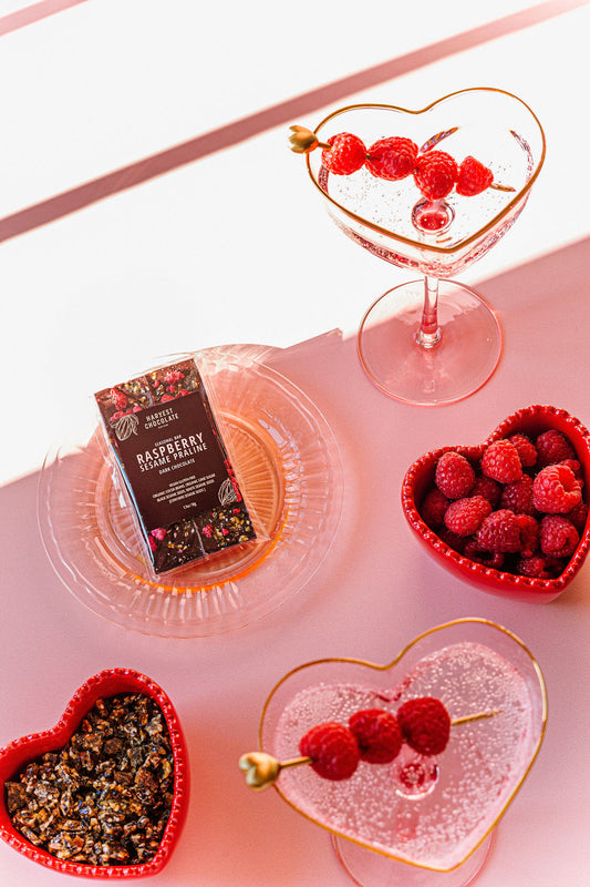 Heart-shaped dishes with fresh raspberries and a nut mix sit beside two martini glasses garnished with raspberry skewers. A clear plate in the center holds a Raspberry Sesame Praline chocolate bar by Harvest Chocolate on a pink surface.