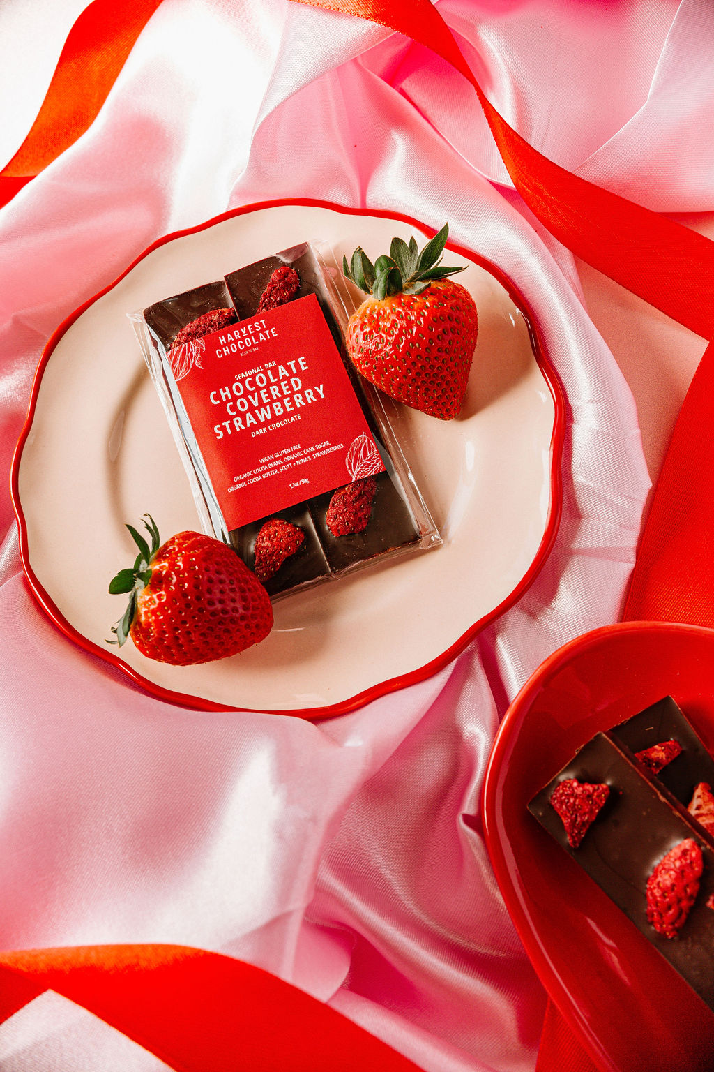 A plate displays a Harvest Chocolate bar, "Chocolate Covered Strawberry," with two fresh strawberries. Red and pink ribbons and fabric enhance the background, featuring a small red dish with dark chocolate pieces infused with Ugandan cocoa beans and strawberry bits.