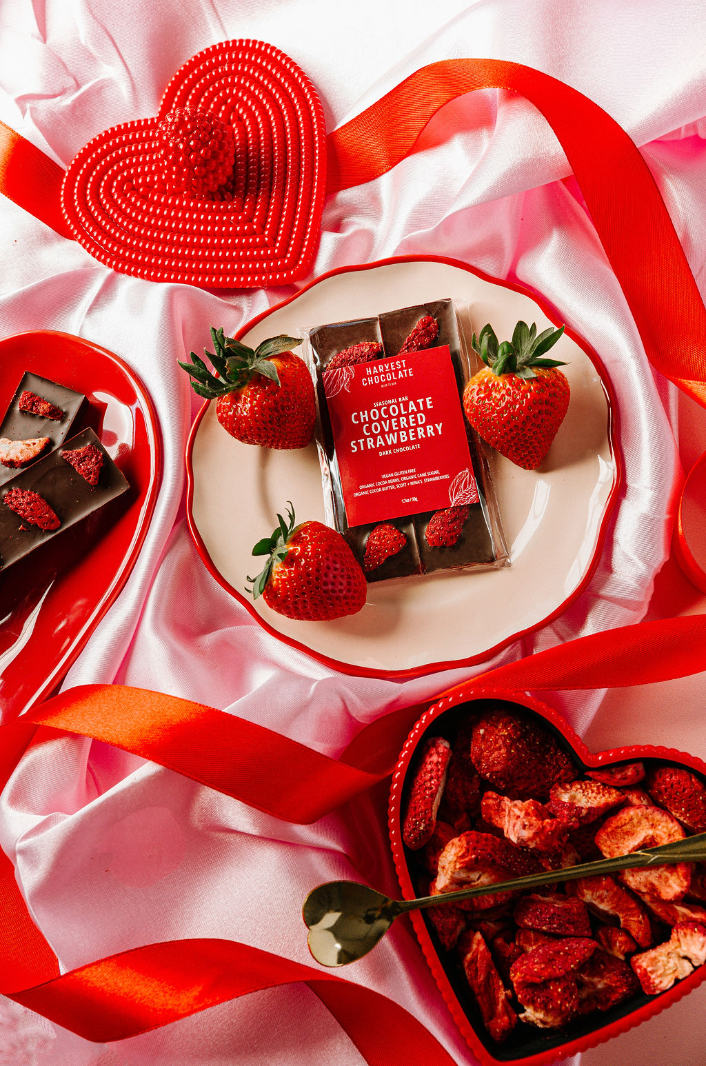 A "Chocolate Covered Strawberry" bar by Harvest Chocolate sits on a white plate adorned with Ugandan cocoa beans, fresh strawberries, and dried slices. It's surrounded by red ribbon, pink satin fabric, and accented with a nearby red heart decoration.