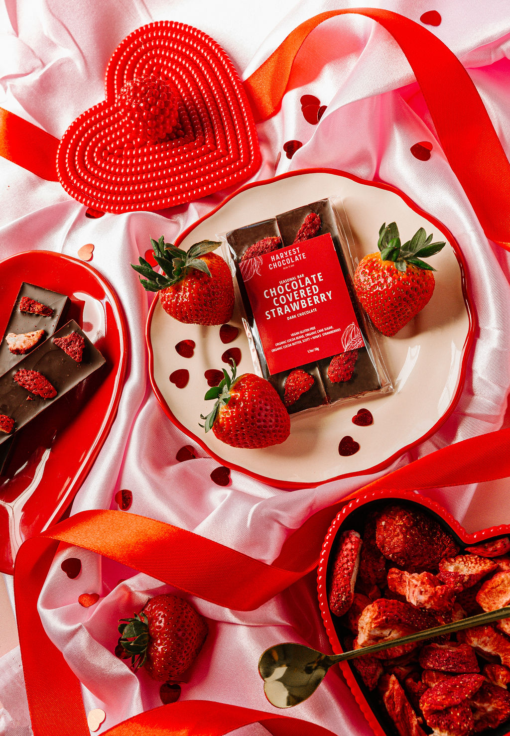 A beautifully arranged display showcases a Chocolate Covered Strawberry bar from Harvest Chocolate, made with rich Ugandan cocoa beans, on a plate encircled by fresh strawberries, chocolate pieces, and red heart decorations. All elements rest elegantly on white satin cloth adorned with red ribbons.