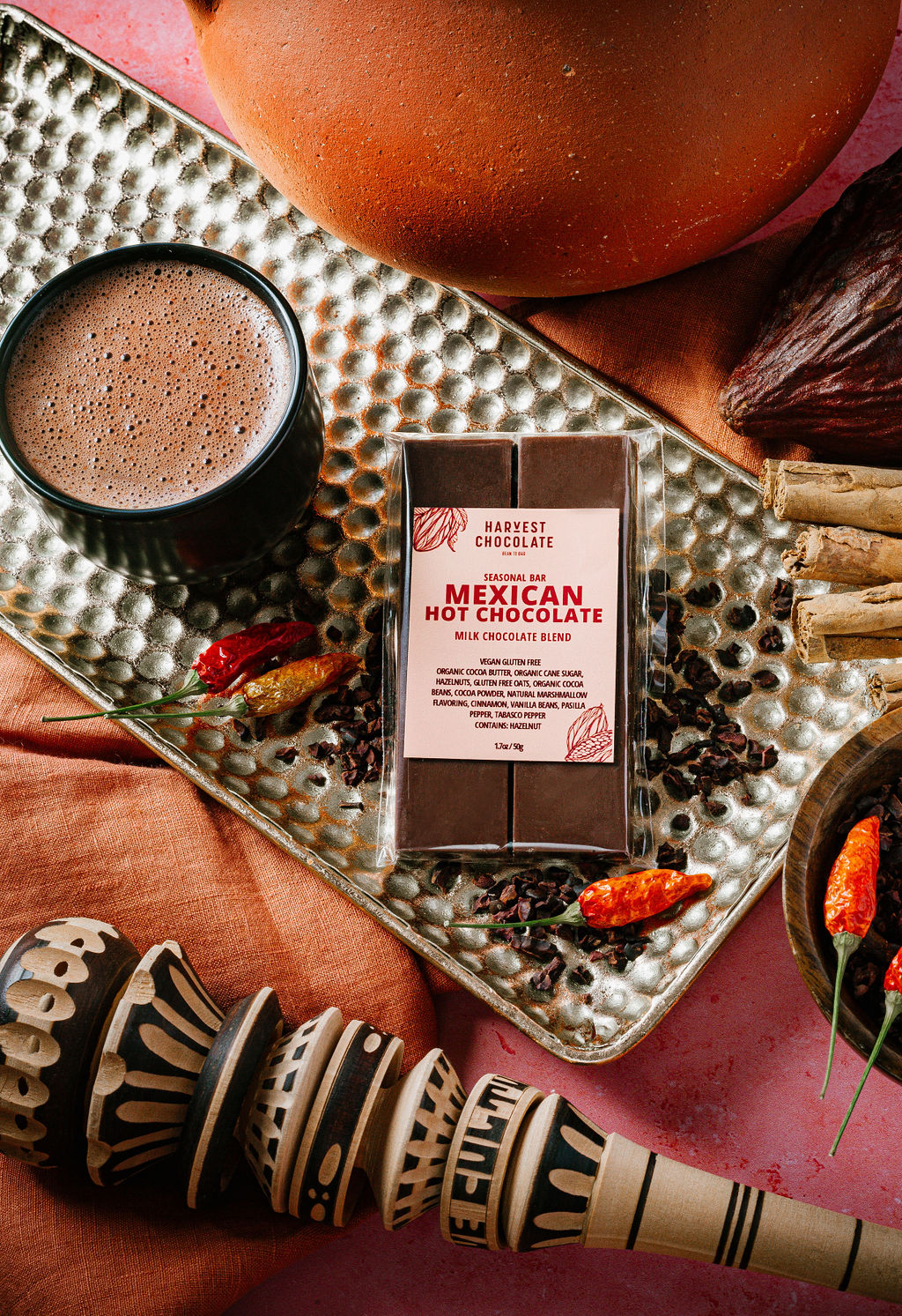 A bar of Harvest Chocolate's Mexican Hot Chocolate, gluten-free and rich in flavor, sits on a textured tray beside a mug of hot chocolate. Nearby are small red chilies and a wooden whisk. Vibrant fabric and scattered cocoa beans complete this inviting scene.