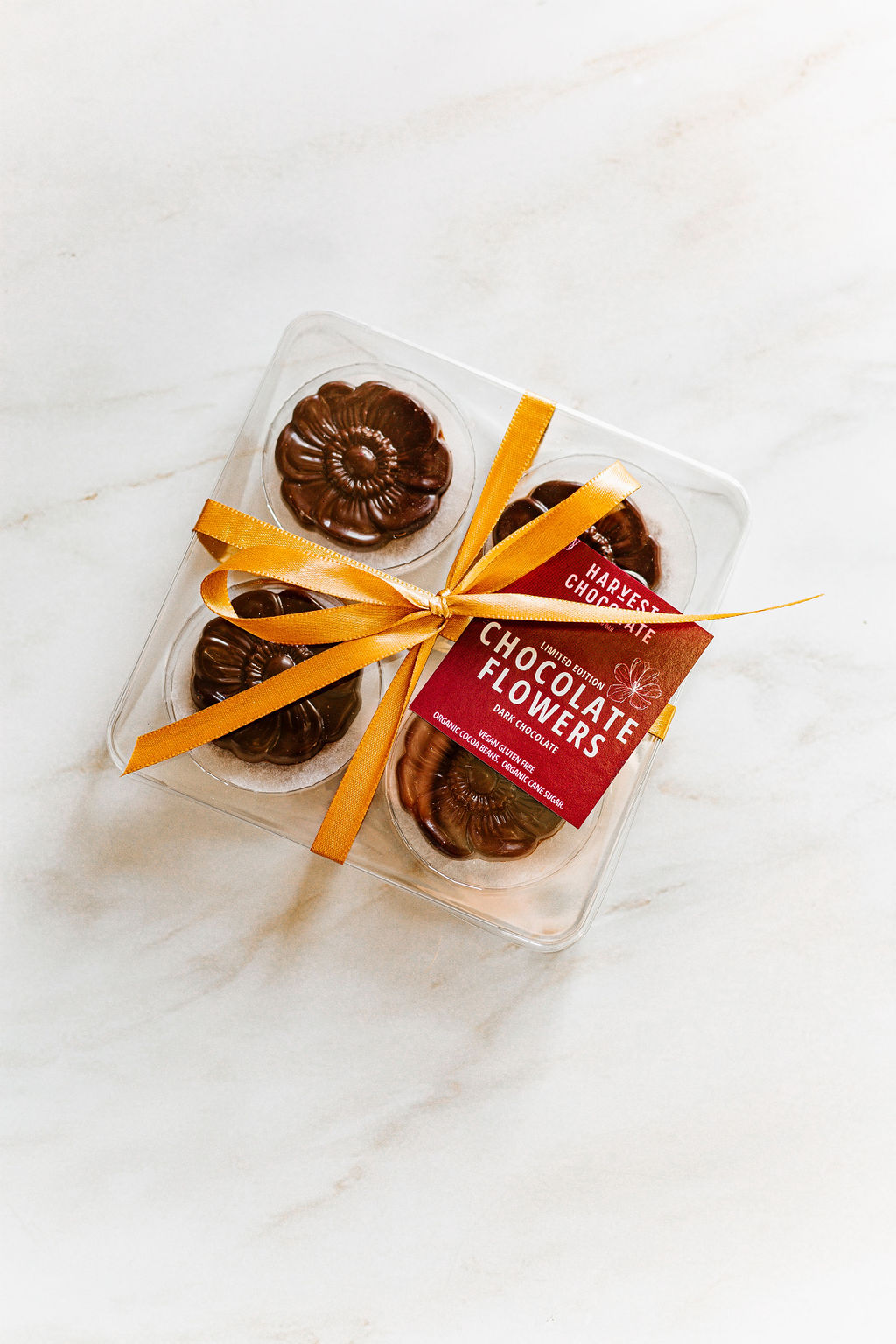 A clear plastic container holds six vegan gluten-free chocolate flowers, secured with an orange ribbon. The label reads "Harvest Chocolate Harvest Chocolate Flowers." Crafted with Ecuadorian cocoa beans, they rest beautifully against a light marbled surface.