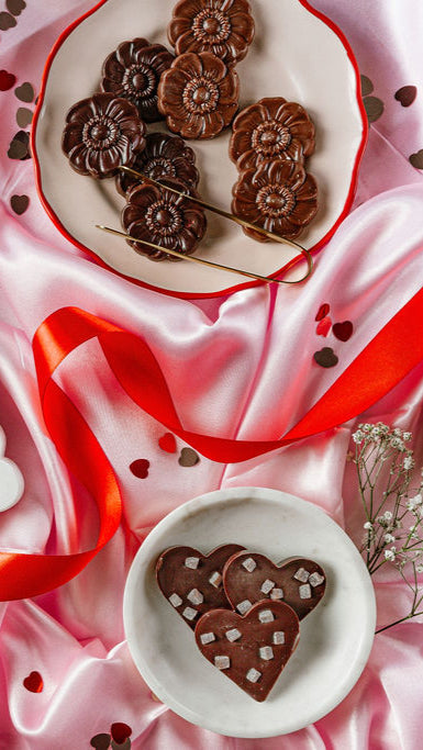 Cherry Jam Hearts cookies by Harvest Chocolate, shaped like flowers and hearts with cherry bits, rest atop pink satin. A red ribbon winds through the scene, surrounded by small heart confetti. White flowers peek in from the lower right corner.