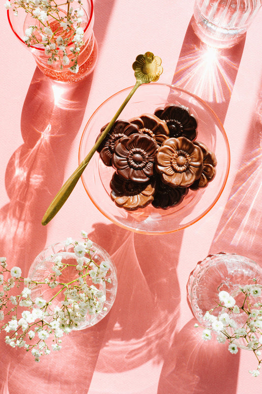 The Harvest Chocolate Flowers, made from exquisite Ecuadorian cocoa by Harvest Chocolate, are beautifully arranged in a glass bowl on a pink surface with a gold spoon. Surrounded by baby's breath and glassware, they cast elegant shadows in the sunlight.