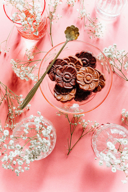 A pink table setting features a glass dish of Harvest Chocolate Flowers, made with rich Ecuadorian cocoa, next to a floral-handled spoon. Delicate white flowers and clear glassware are elegantly arranged around the dish by Harvest Chocolate.